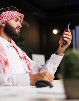 middle-eastern-businessman-dressed-traditionally-sits-holds-his-smartphone-desk-his-office-arab-guy-grasping-mobile-device-using-desktop-computer-nearby (1)