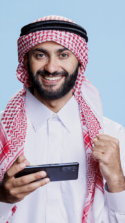 Happy muslim man playing online game on smartphone and showing winner concept studio portrait. Arab person winning videogame and looking at camera with clenched fist and excited expression